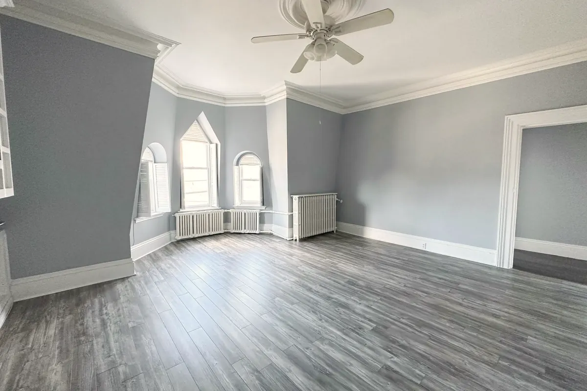 A large room with grey walls, new plank floors, a ceiling fan and arched windows in a bay.